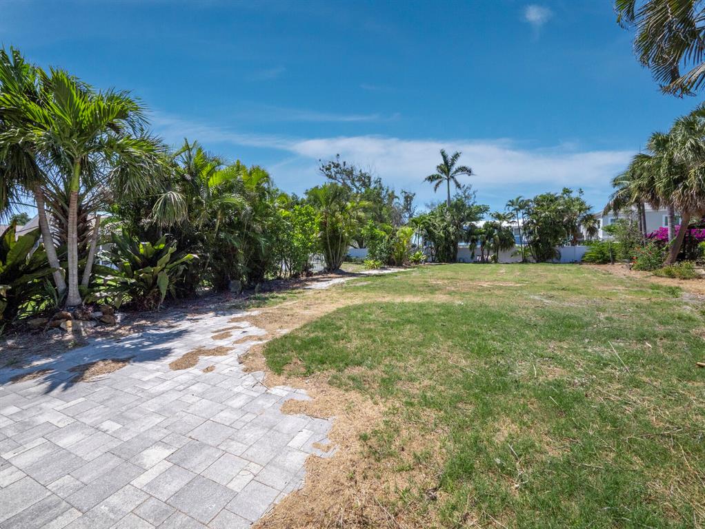 A paved patio area transitions into a large, grassy backyard surrounded by various tropical trees — 450 FIREHOUSE COURT, LONGBOAT KEY, FL