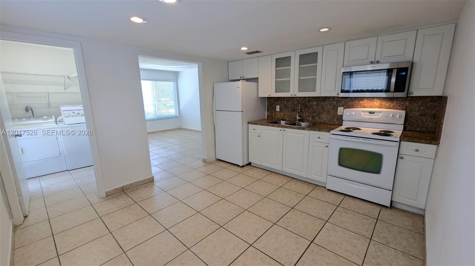 A kitchen with white cabinets, white appliances, and tan tile flooring leads to a laundry room — 3525 SW 25th Ter # 1, Miami FL 33133, MIAMI, FL