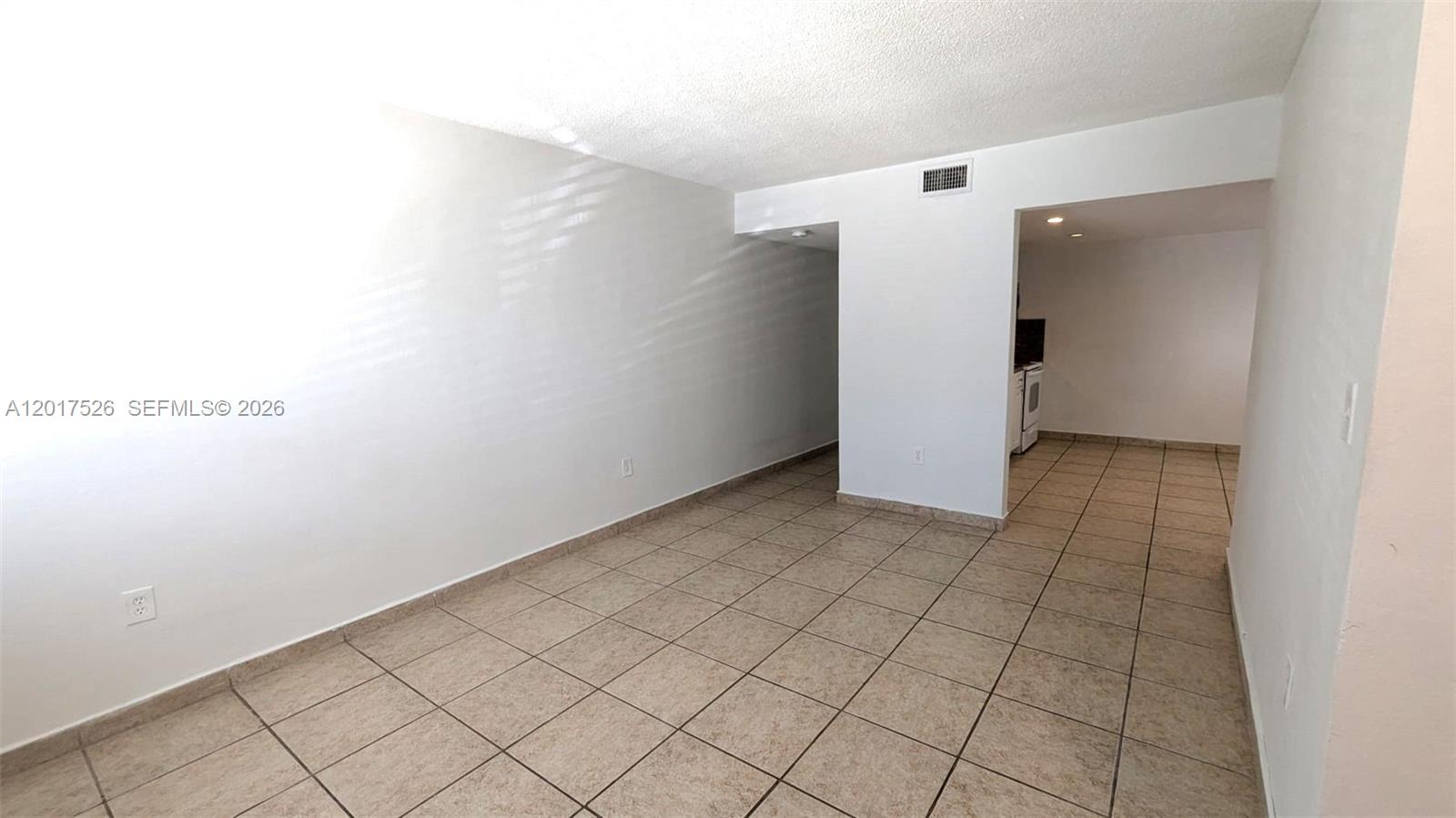 An empty room with beige tile flooring and white walls leads into a kitchen area — 3525 SW 25th Ter # 1, Miami FL 33133, MIAMI, FL
