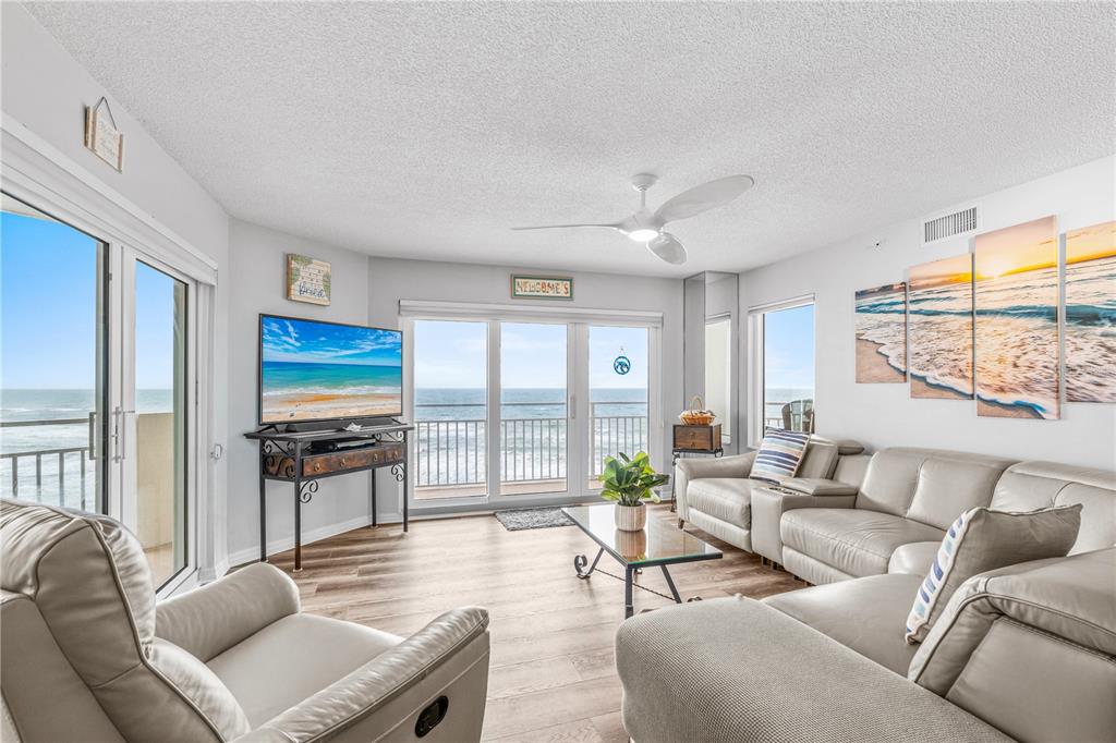 A living room features light-colored leather furniture, wood-look flooring, and sliding glass doors overlooking the ocean — 2967 S ATLANTIC AVENUE UNIT 903, DAYTONA BEACH SHORES, FL