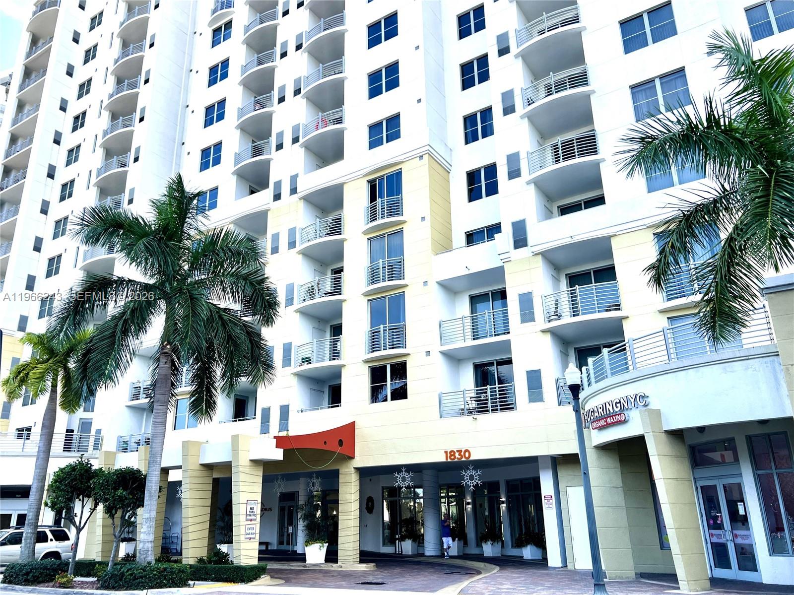 The exterior of a multi-story apartment building features balconies, palm trees, and a ground-level storefront — 1830 Radius Dr # 1203, Hollywood FL 33020, HOLLYWOOD, FL