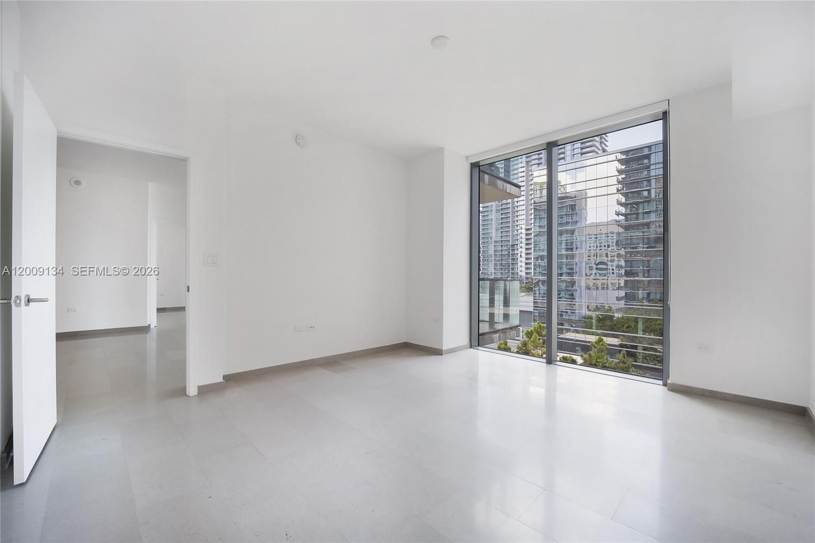 An empty room with white walls, light tile flooring, and floor-to-ceiling windows overlooking city buildings — 88 SW 7th St # 1009, Miami FL 33130, MIAMI, FL