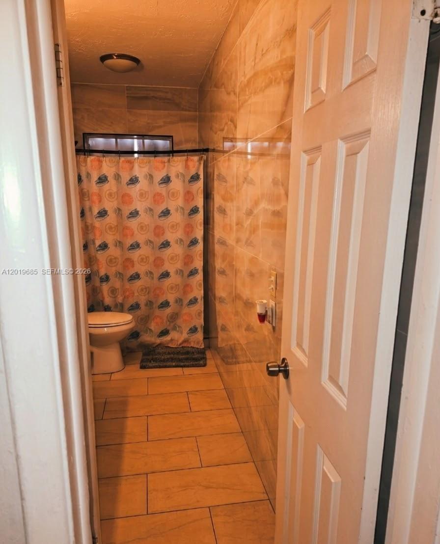 A narrow bathroom features tan tiled walls and flooring with a patterned shower curtain — 571 NE 172nd St, North Miami Beach FL 33162, NORTH MIAMI BEACH, FL