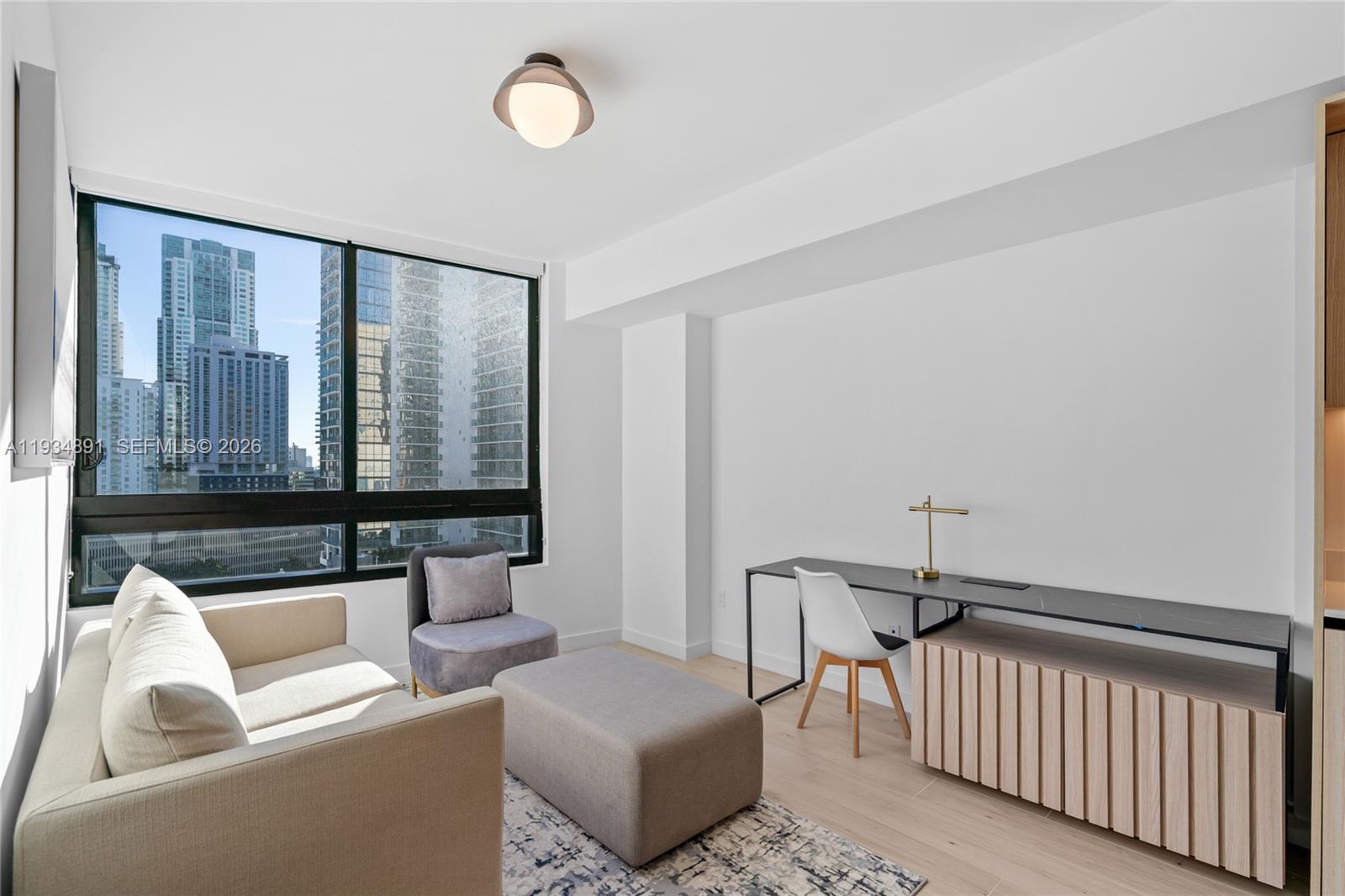 A modern room features a sofa, desk, and large windows overlooking a city skyline — 225 N Miami Ave # 1405, Miami FL 33132, MIAMI, FL