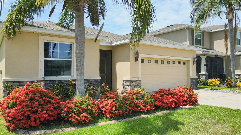 A single-story house with a tan stucco exterior, a two-car garage, and red flowering bushes — 417 BLAKELY COURT, RUSKIN, FL