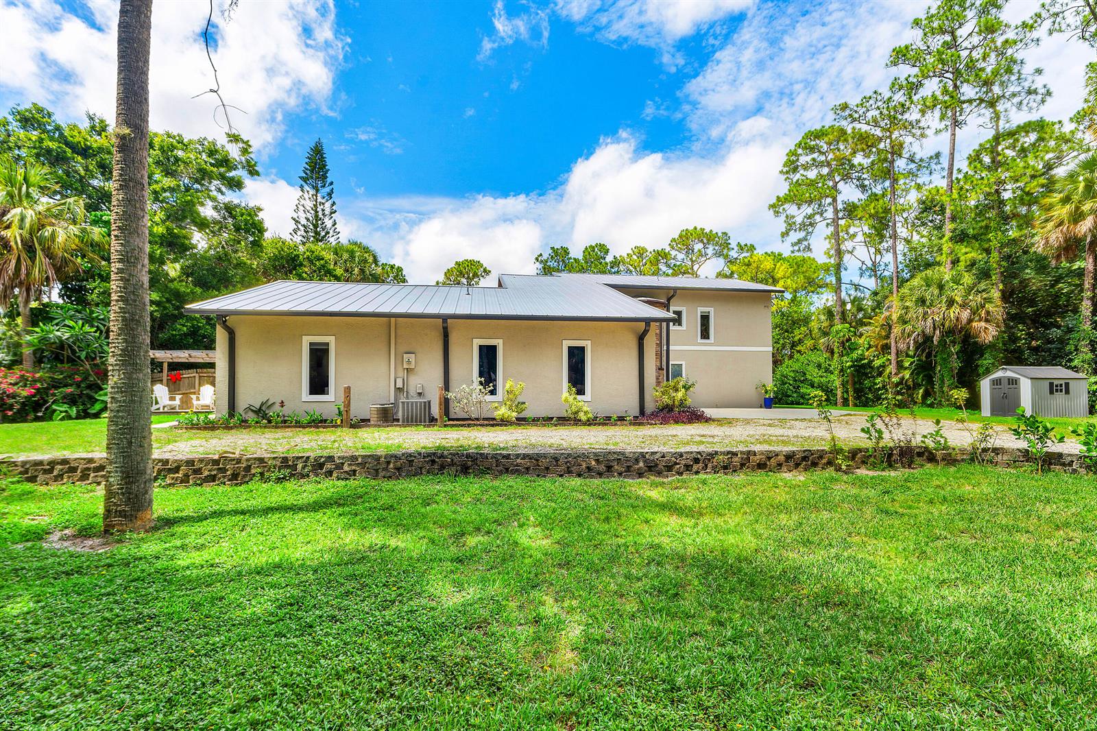 A beige house with a metal roof sits behind a stone retaining wall and lawn — 9743 169th Court N, Jupiter FL 33478, JUPITER, FL