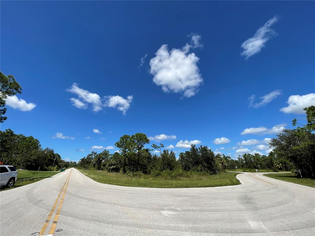 A paved road curves through a grassy, wooded area under a bright blue sky — 27554 MONTE CRISTO BOULEVARD, PUNTA GORDA, FL
