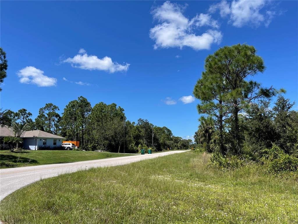 A paved road runs past a grassy lot with trees and a house in the distance — 27554 MONTE CRISTO BOULEVARD, PUNTA GORDA, FL