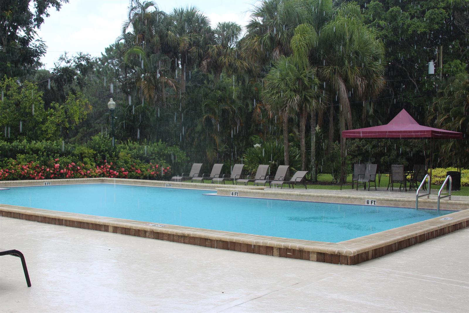 A rectangular swimming pool with depth markers sits on a concrete deck during rain — 7705 Forest Green Lane, Boynton Beach FL 33436, BOYNTON BEACH, FL