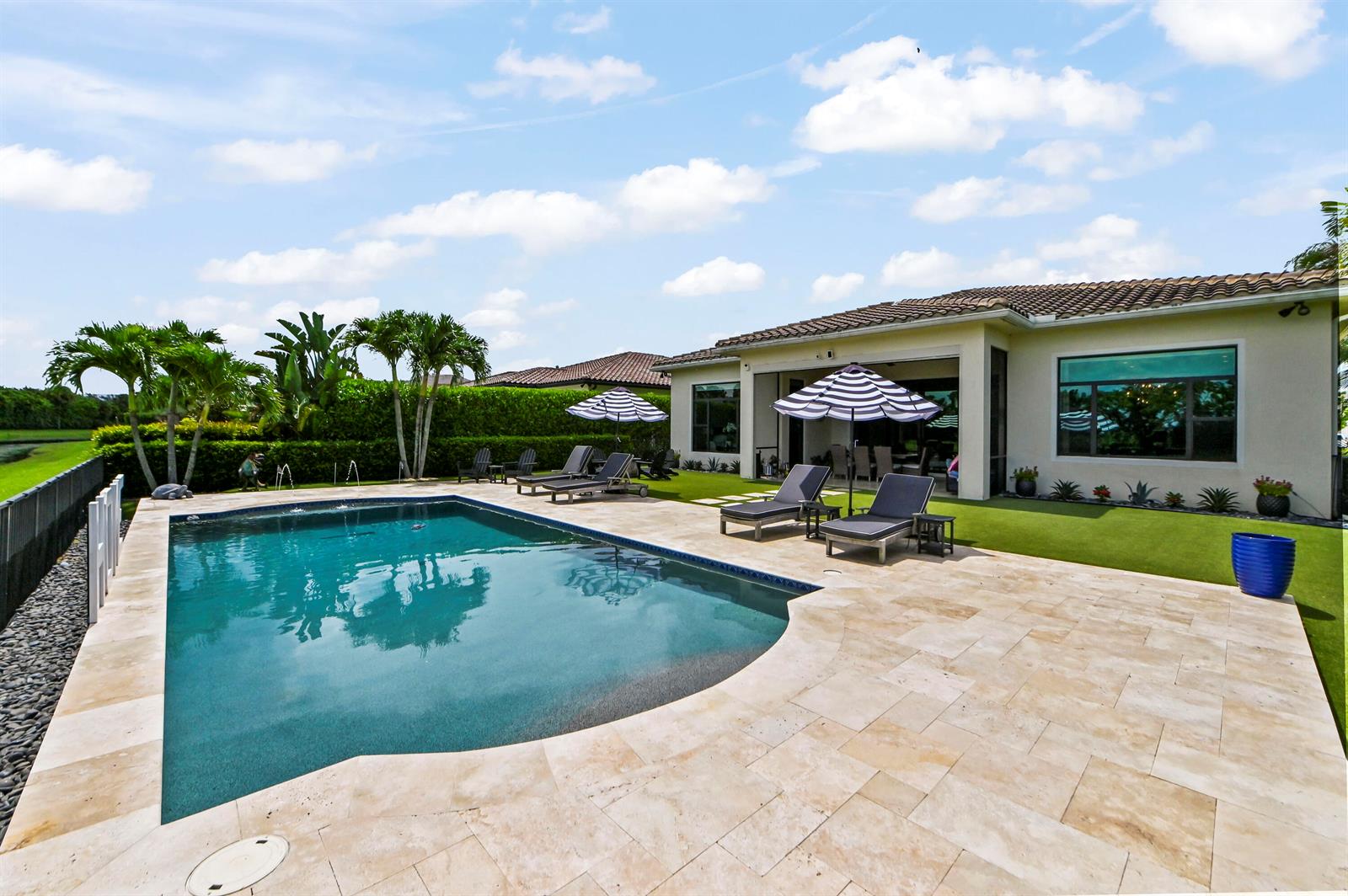 A backyard features a swimming pool, stone patio, lounge chairs, umbrellas, and a house exterior — 9307 Grand Prix Lane, Boynton Beach FL 33472, BOYNTON BEACH, FL