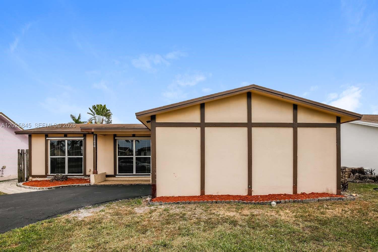 A single-story house with tan stucco walls, brown trim, and a dark asphalt driveway — SUNRISE, FL