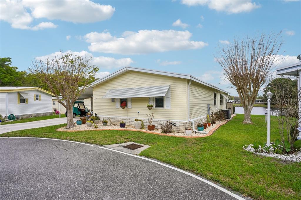 A light yellow manufactured home with a carport and landscaped garden sits on a corner lot — 122 TAMARISK WAY, LEESBURG, FL