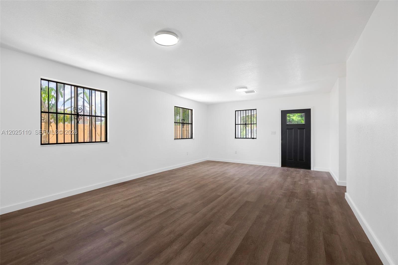 An empty room features white walls, dark wood-look flooring, barred windows, and a black door — 10204 SW 173rd St, Miami FL 33157, MIAMI, FL