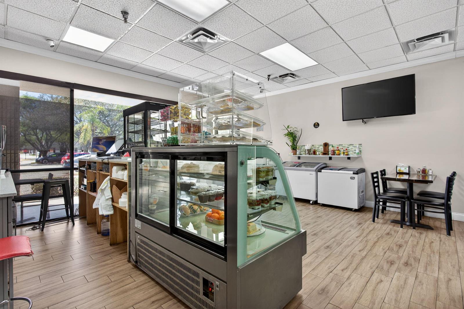 A commercial cafe interior features a refrigerated display case, wood-look flooring, and dining tables — Null W Null, Coconut Creek FL 33073, COCONUT CREEK, FL