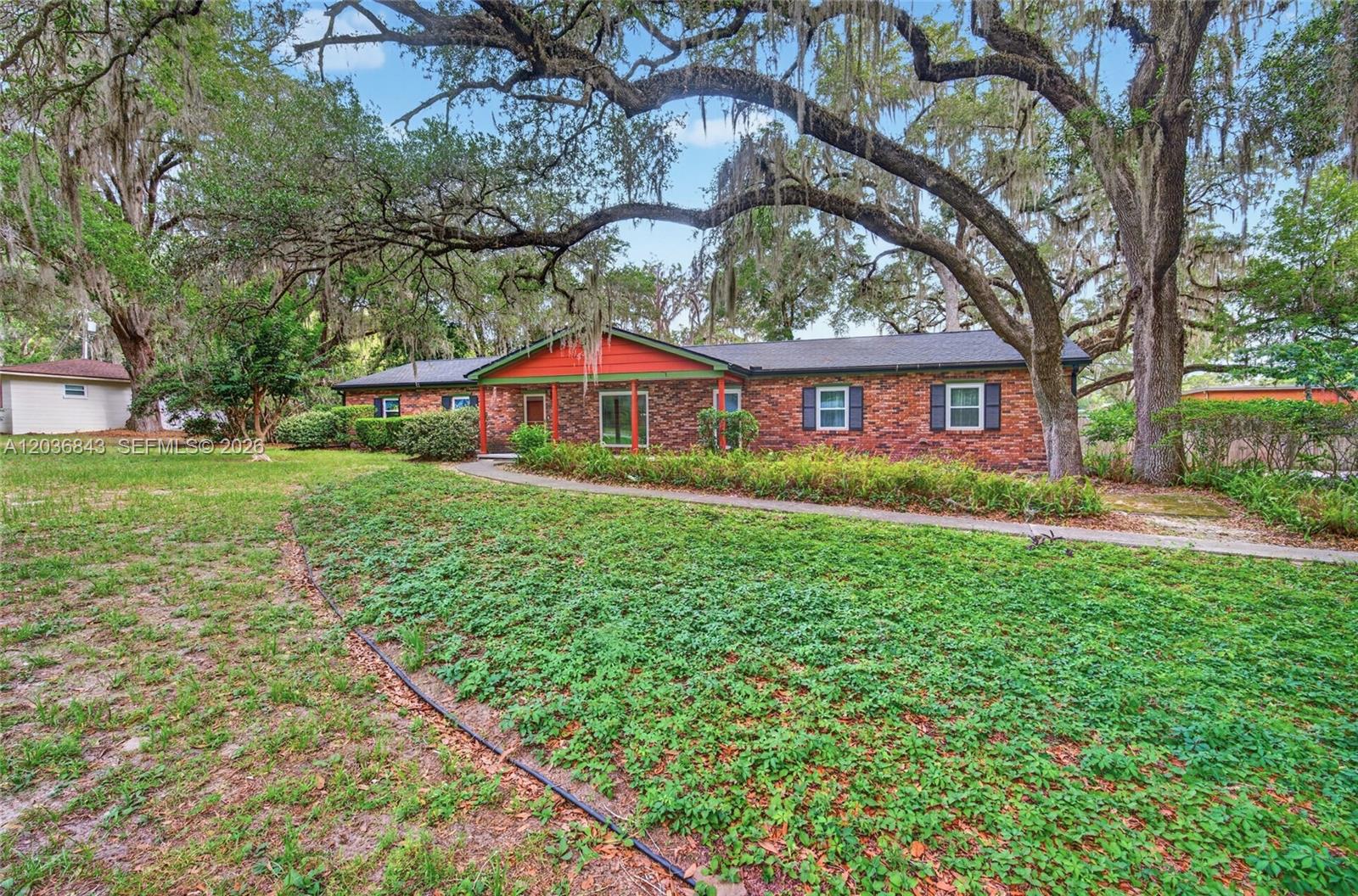 A single-story brick house with a red-trimmed porch sits under large, moss-covered oak trees — 1944 SW 48th Avenue, Gainesville FL 32608, GAINESVILLE, FL
