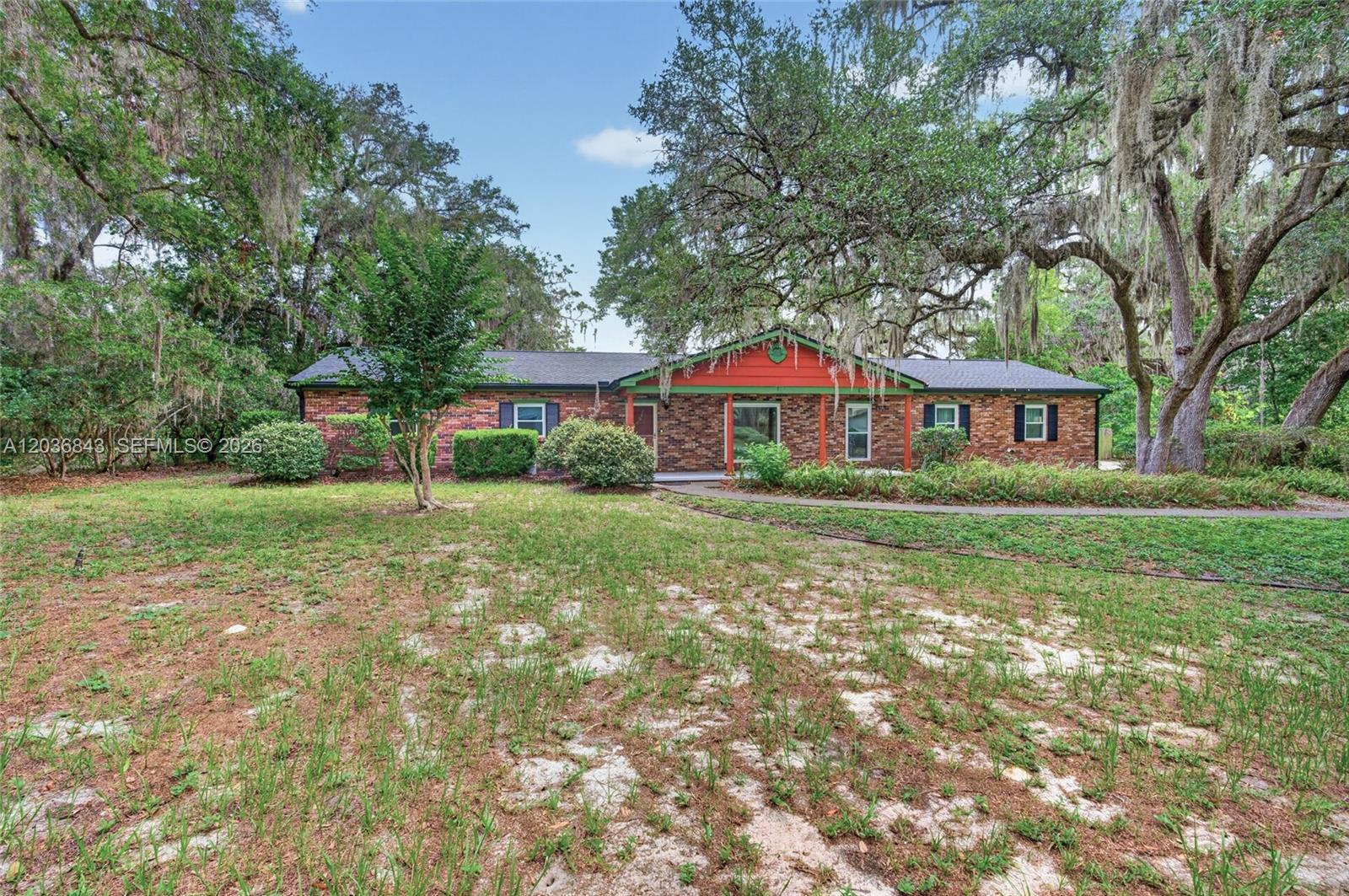 A single-story brick house with a red-painted porch sits on a grassy, wooded lot — 1944 SW 48th Avenue, Gainesville FL 32608, GAINESVILLE, FL