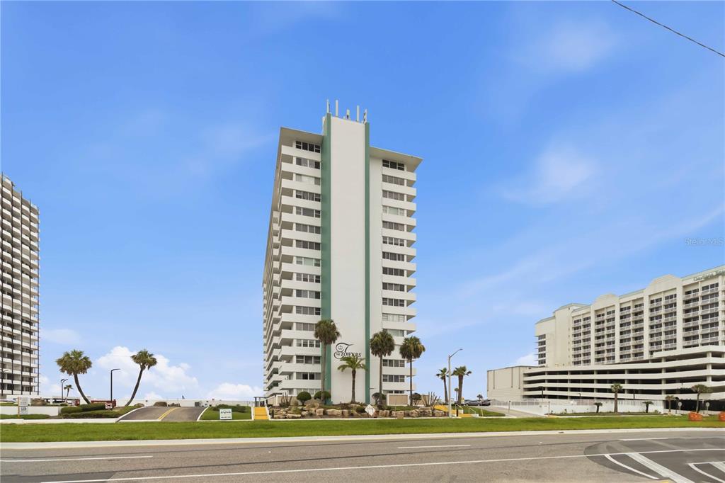 A tall white residential tower stands against a blue sky near a paved road — 2800 N ATLANTIC AVENUE UNIT 1105, DAYTONA BEACH, FL