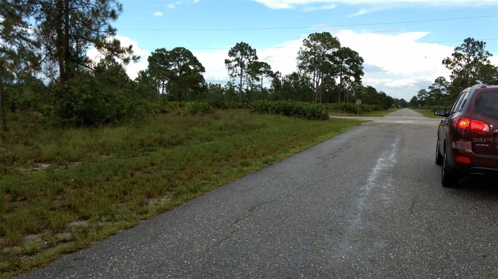 A paved road leads toward a wooded area with a parked car on the right — 4006 E 5TH STREET, LEHIGH ACRES, FL
