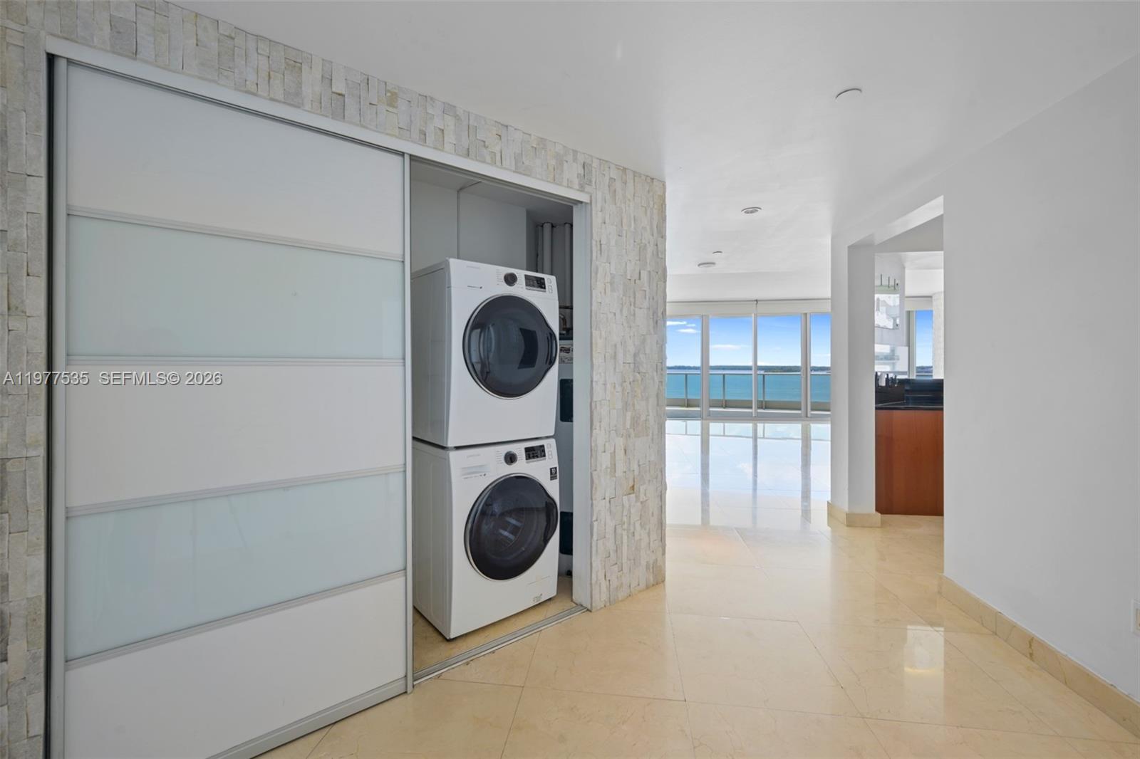 A stacked washer and dryer unit sits in a closet next to a bright living area — 1331 Brickell Bay Dr # 911, Miami FL 33131, MIAMI, FL