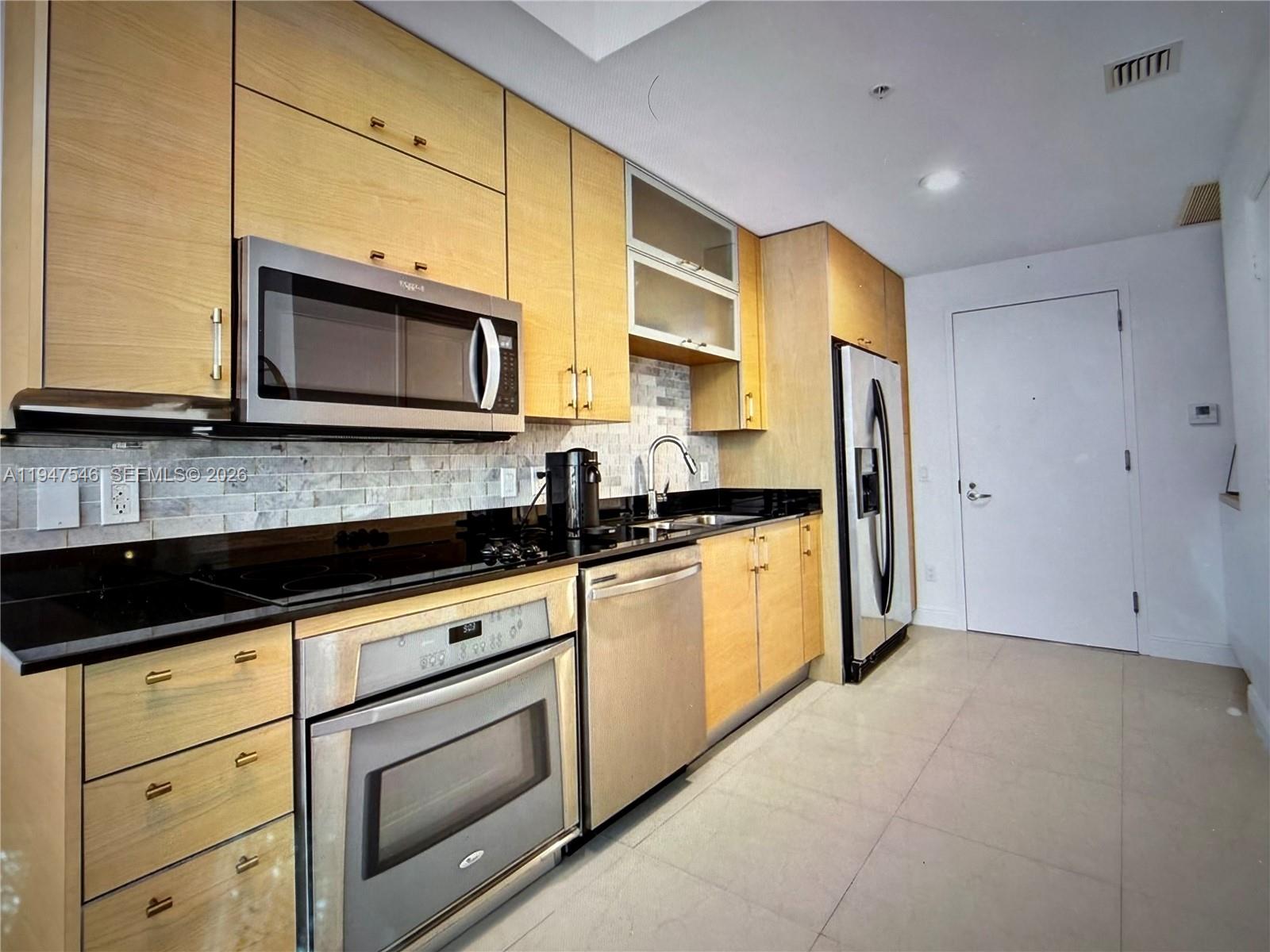 A kitchen features light wood cabinets, black countertops, stainless steel appliances, and a tiled backsplash — 60 SW 13th St # 3214, Miami FL 33130, MIAMI, FL