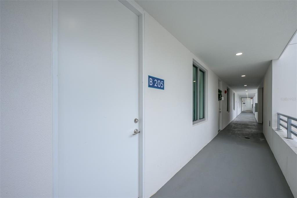 A white door labeled B 205 opens onto a long, covered outdoor apartment building hallway — 1703 N TAMIAMI TRAIL UNIT 205, SARASOTA, FL