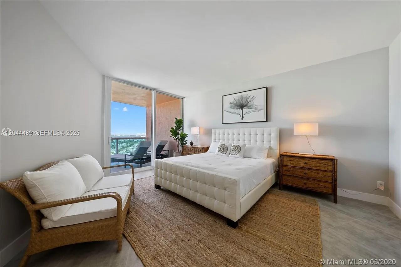 A bedroom features a tufted white bed, a wicker loveseat, and a balcony with ocean views — 300 S Pointe Dr # 3604, Miami Beach FL 33139, MIAMI BEACH, FL
