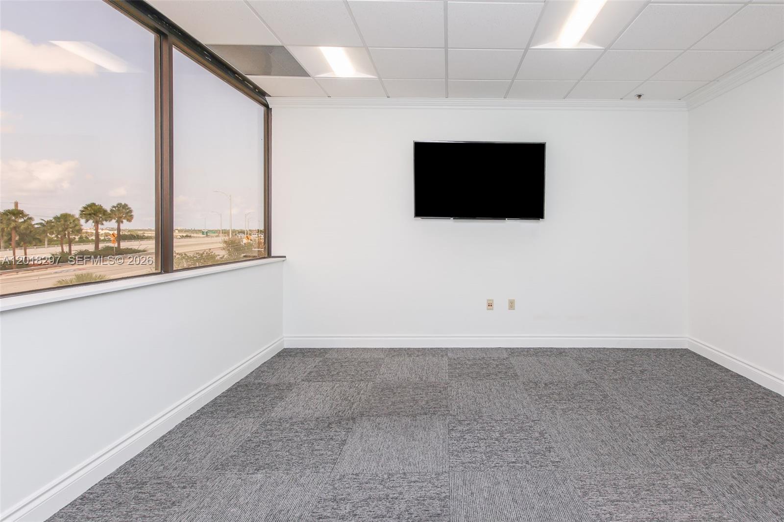 An empty office with gray carpet, white walls, a wall-mounted television, and a large window — 1900 Glades Rd # 301, Boca Raton FL 33431, BOCA RATON, FL