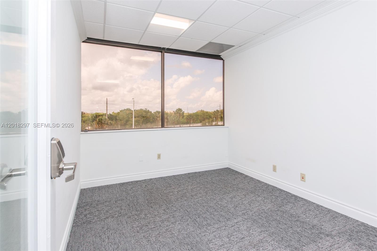 An empty office features gray carpet, white walls, a drop ceiling, and a large window — 1900 Glades Rd # 301, Boca Raton FL 33431, BOCA RATON, FL