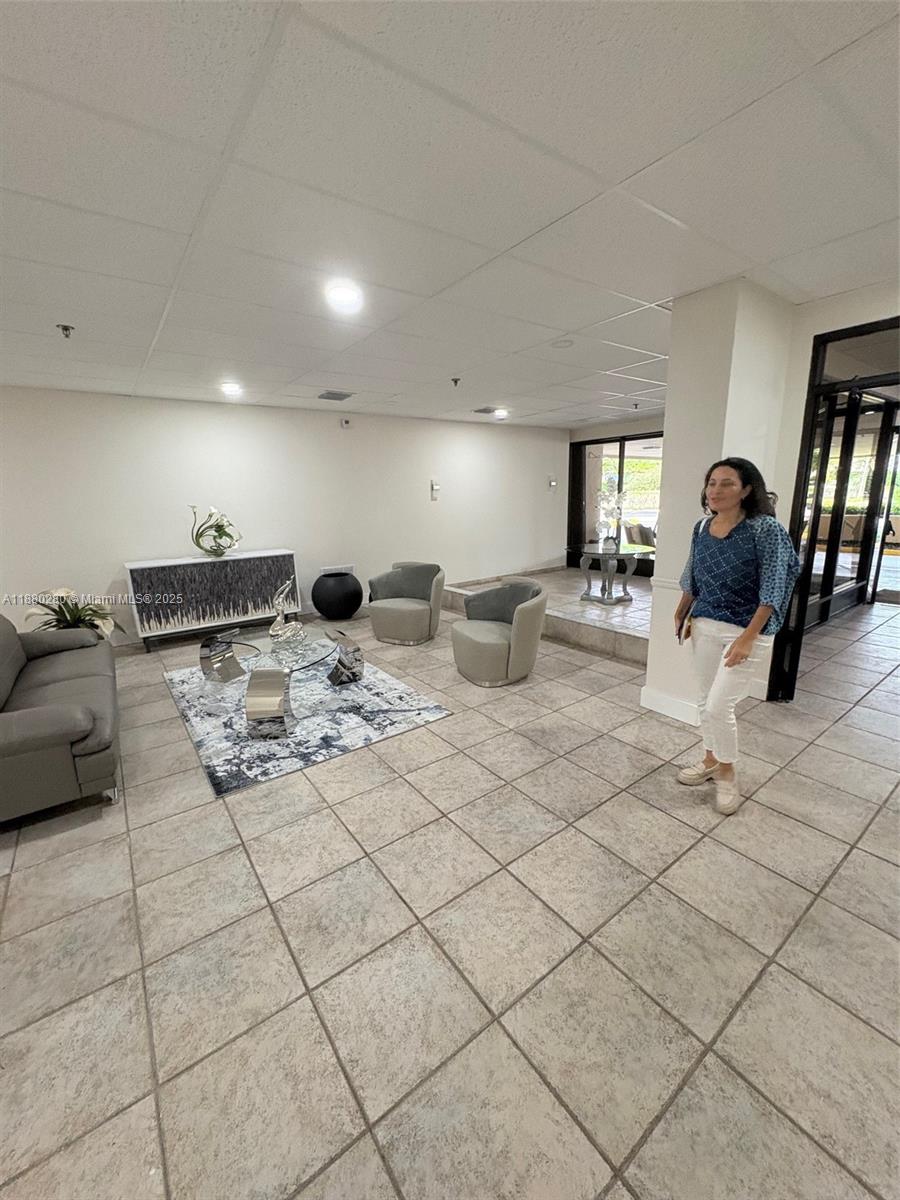 A woman stands in a tiled lobby featuring a seating area with armchairs and furniture — 900 NE 195th St # 702, Miami FL 33179, MIAMI, FL