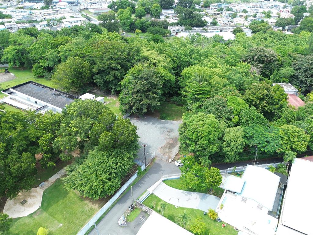An aerial view shows a gravel lot surrounded by dense trees and nearby buildings — SECTOR LA ALDEA, BAYAMON, PR