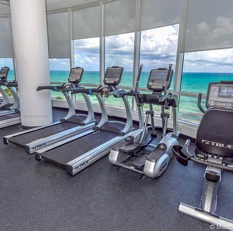 A gym features several treadmills and an elliptical machine facing floor-to-ceiling windows overlooking the ocean — 6365 Collins Ave # 904, Miami Beach FL 33141, MIAMI BEACH, FL