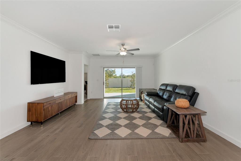 A living room features wood-look tile flooring, a black leather sofa, and a sliding glass door — 17322 HOLLY WELL AVENUE, WIMAUMA, FL