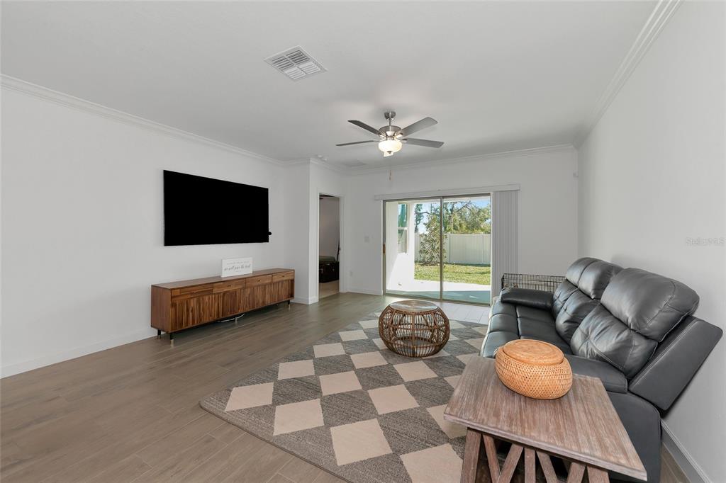 A living room features light wood-look flooring, a black leather sofa, and a sliding glass door — 17322 HOLLY WELL AVENUE, WIMAUMA, FL