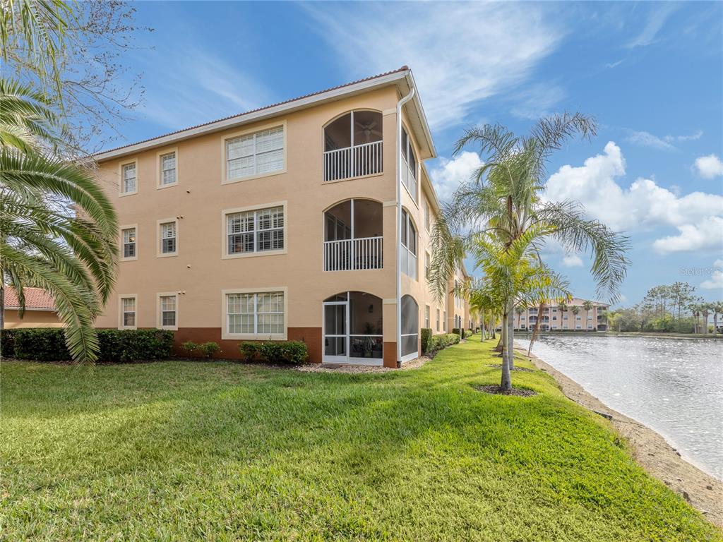 A three-story apartment building with screened balconies sits beside a pond and palm trees — 1731 AUBURN LAKES DRIVE UNIT 24, VENICE, FL