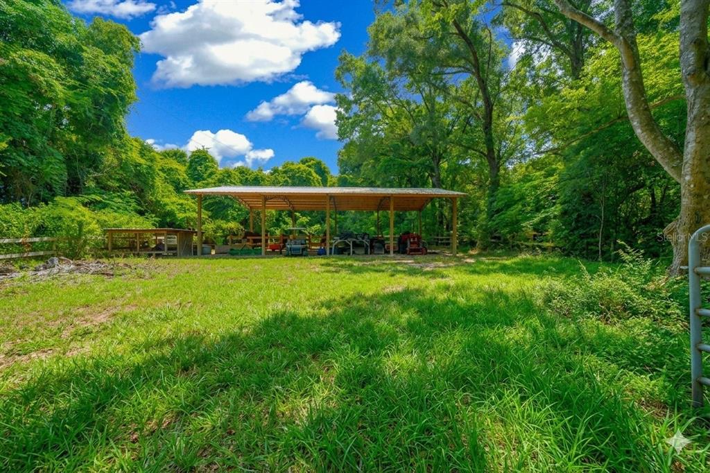 A large open-sided metal pole barn stands in a grassy field surrounded by dense trees — 13576 NW COUNTY ROAD 235, ALACHUA, FL