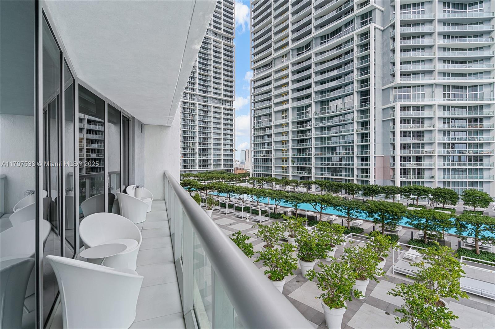 A balcony with white chairs overlooks a swimming pool and landscaped courtyard between high-rise buildings — 495 Brickell Ave # 1702, Miami FL 33131, MIAMI, FL