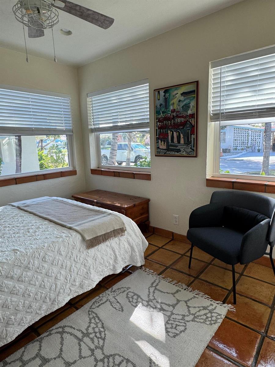 A bedroom with a bed, a dark armchair, tile floors, and windows overlooking a street — 256 E 24th Street, Riviera Beach FL 33404, RIVIERA BEACH, FL