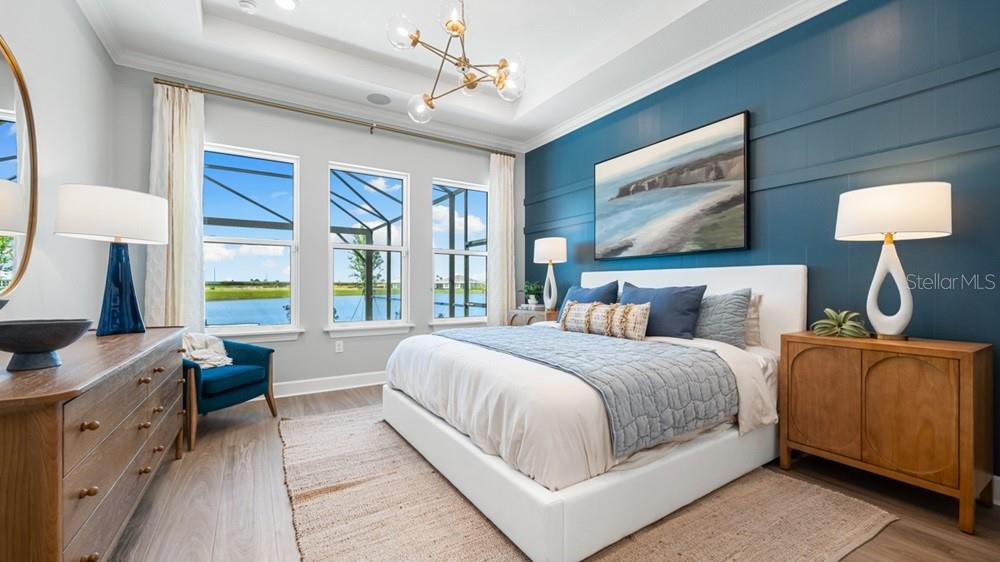 A bedroom features a blue accent wall, a white bed, and windows overlooking a lake — 7420 MOONBEAM AVENUE, SARASOTA, FL