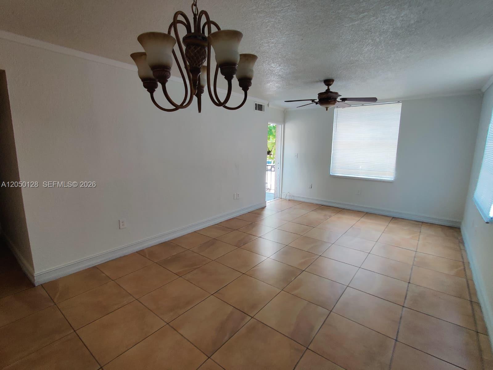 An empty room features tan tile flooring, a chandelier, a ceiling fan, and white walls — 5705 W 20th Ave # 215, Hialeah FL 33012, HIALEAH, FL