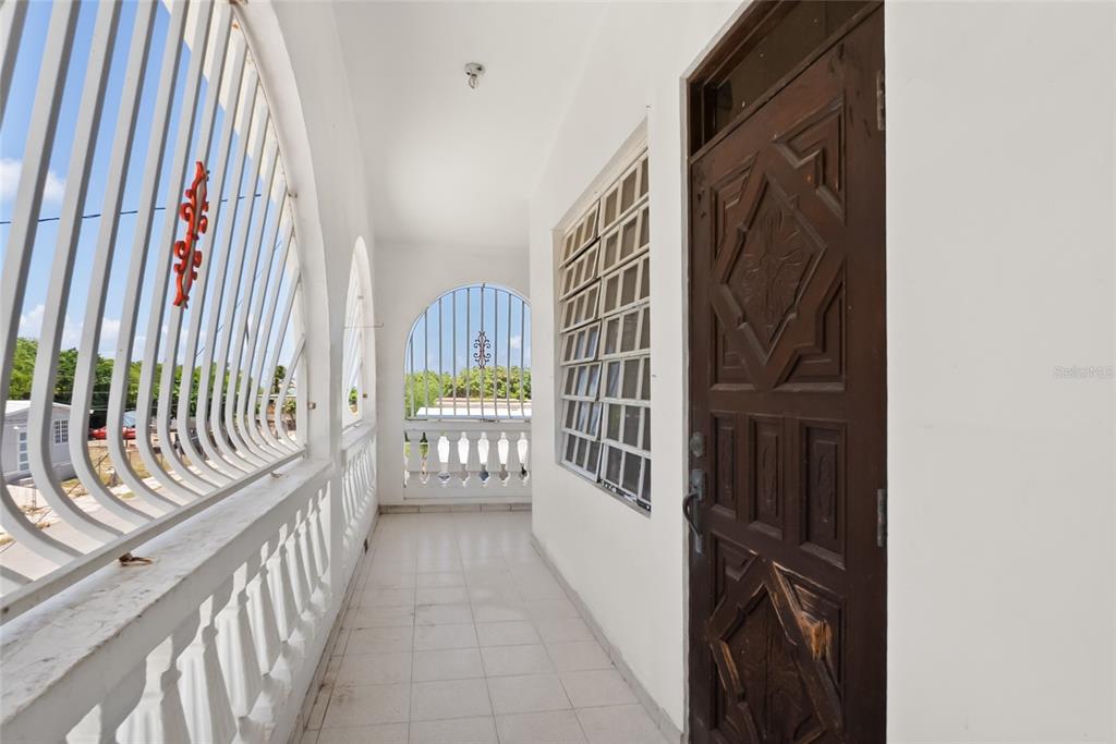 A tiled balcony features white arched railings, a dark wooden door, and louvered windows — 638 CALLE B COM. SERRANO, JUANA DIAZ, PR
