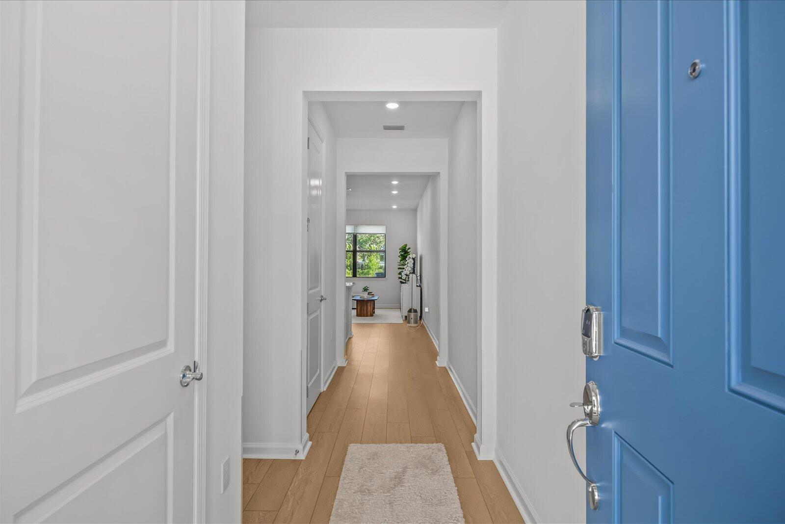 A bright hallway with light wood floors leads from a blue front door toward windows — 276 SW Sally Way, Stuart FL 34997, STUART, FL