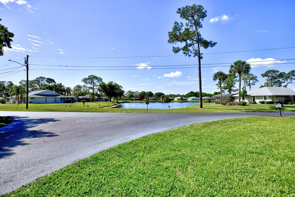A paved road curves past a grassy lawn toward a pond and residential houses — 3601 DIVOT ROAD, SEBRING, FL