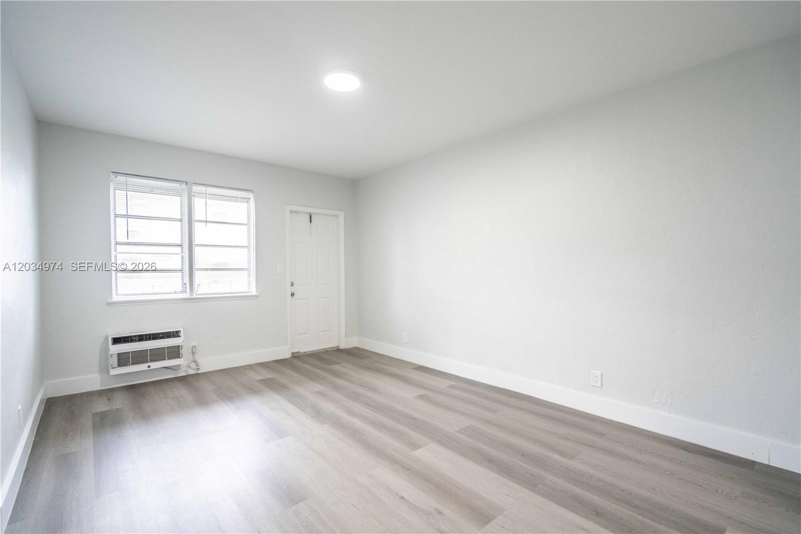 An empty room features light gray walls, wood-look flooring, a window, and a wall-mounted air conditioner — 1208 71st St # 3, Miami Beach FL 33141, MIAMI BEACH, FL