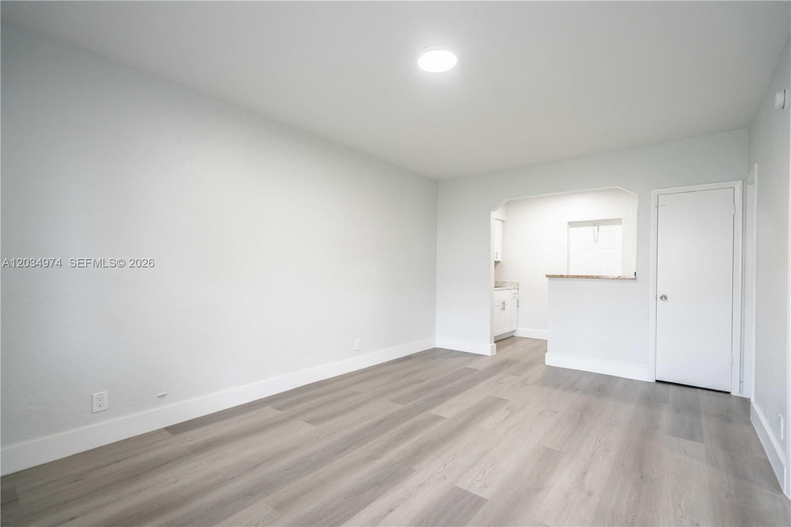 An empty room with light gray walls, wood-look flooring, and a doorway to a kitchen — 1208 71st St # 3, Miami Beach FL 33141, MIAMI BEACH, FL