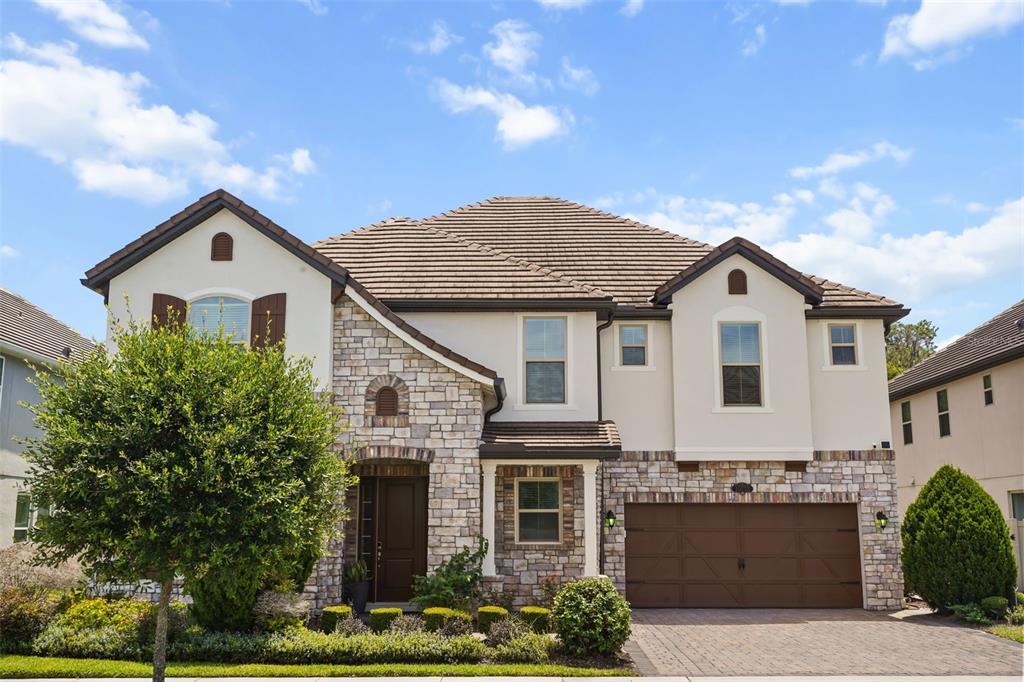 A two-story house features a stone and stucco facade, a brown garage, and a tiled roof — 10713 BELLE MAISONS DRIVE, ORLANDO, FL