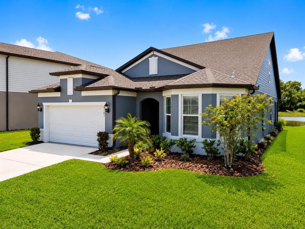 A single-story gray house with a white garage door and a landscaped front yard — 9107 RALLY SPRING LOOP, WESLEY CHAPEL, FL