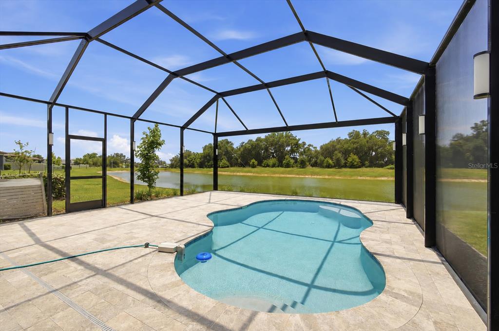 A screened-in pool patio features light stone pavers overlooking a pond and grassy landscape — 9107 RALLY SPRING LOOP, WESLEY CHAPEL, FL