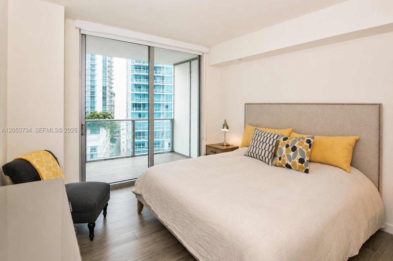 A bedroom features a bed with yellow pillows, a gray armchair, and a balcony view — 1100 S Miami Ave # 1711, Miami FL 33130, MIAMI, FL