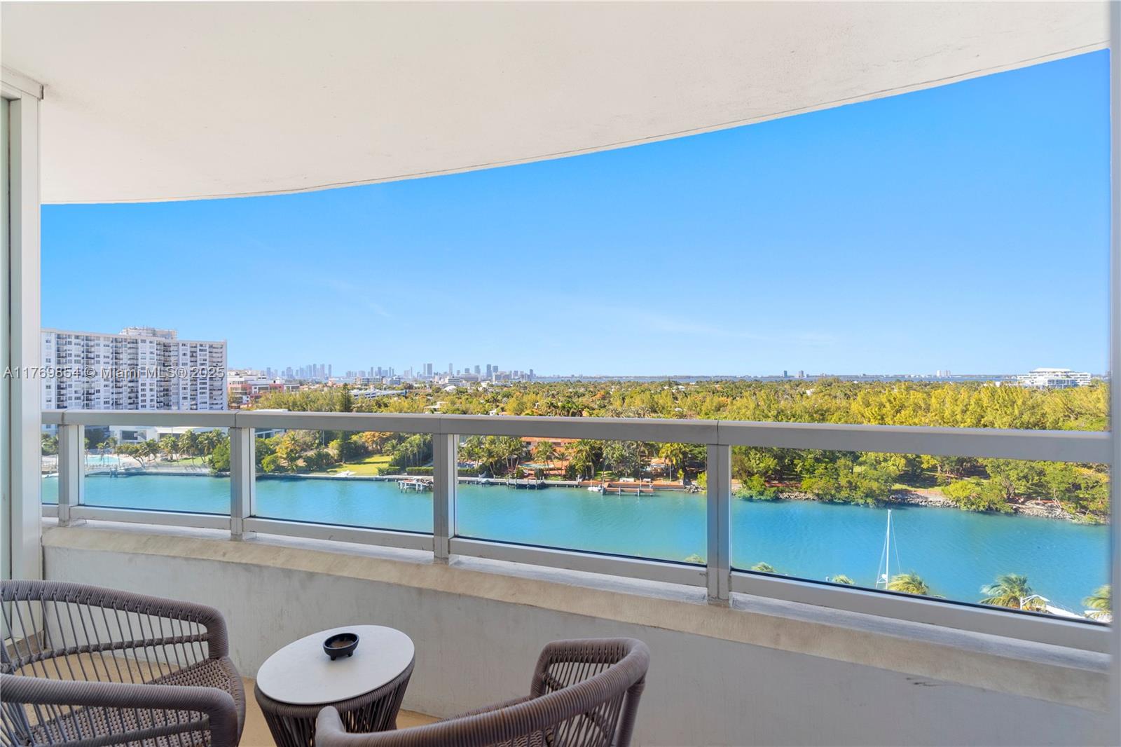 A balcony with two chairs and a small table overlooks a waterway and city skyline — 4401 Collins Ave # 1017, Miami Beach FL 33140, MIAMI BEACH, FL