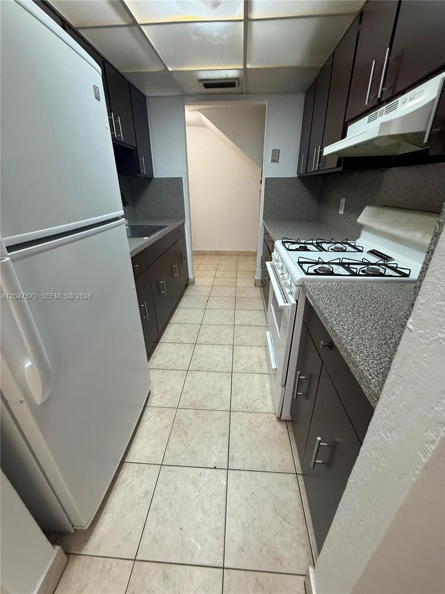 A galley kitchen features dark cabinets, white appliances, speckled countertops, and light-colored tile flooring — 1305 W 53rd St # 406, Hialeah FL 33012, HIALEAH, FL
